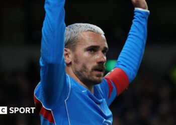 Atletico Madrid forward Antoine Griezmann, with bleached blond hair and facial hair, has both arms raised as he wears blue and red kit