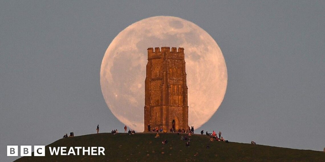A huge pink hued Moon sits in a grey sky, behind the tower on Glastonbury Tor with people milling around at the base