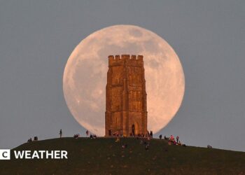 A huge pink hued Moon sits in a grey sky, behind the tower on Glastonbury Tor with people milling around at the base