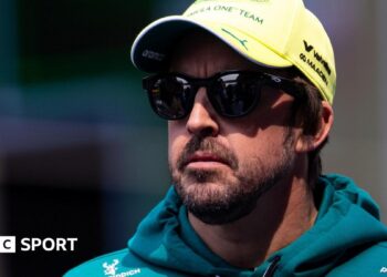 Fernando Alonso wearing sunglasses, an Aston Martin hoodie and a yellow cap