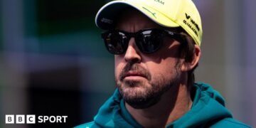 Fernando Alonso wearing sunglasses, an Aston Martin hoodie and a yellow cap