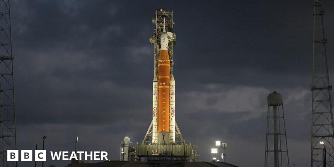 Orange rocket with white tip stands vertically ready to lift off lit up by flood lights