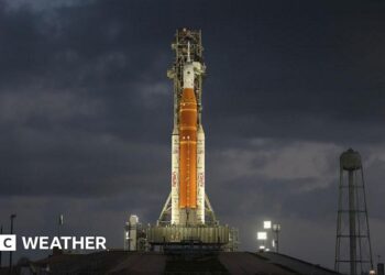 Orange rocket with white tip stands vertically ready to lift off lit up by flood lights