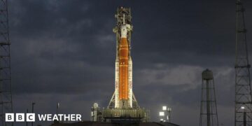 Orange rocket with white tip stands vertically ready to lift off lit up by flood lights