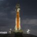 Orange rocket with white tip stands vertically ready to lift off lit up by flood lights