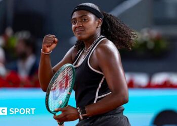 Hailey Baptiste, wearing a black vest and shorts, black headband and a ponytail, holds her green Wilson racket in her left hand and punches her right fist in celebration