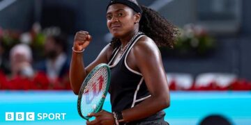Hailey Baptiste, wearing a black vest and shorts, black headband and a ponytail, holds her green Wilson racket in her left hand and punches her right fist in celebration