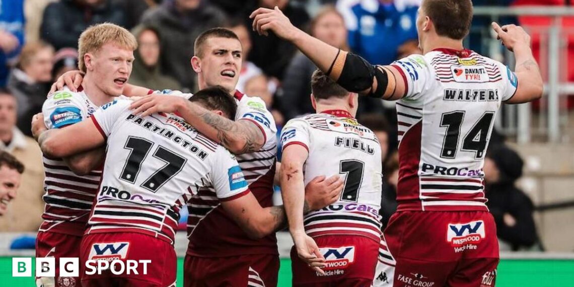 Wakefield's Tom Johnstone diving towards the line to score a try one-handed for his side under pressure from two Wigan defenders in the corner.