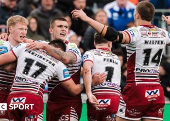 Wakefield's Tom Johnstone diving towards the line to score a try one-handed for his side under pressure from two Wigan defenders in the corner.
