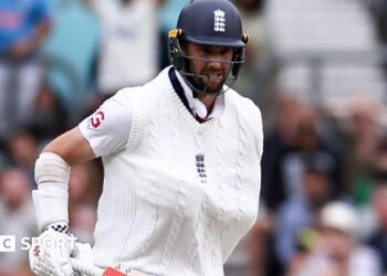 Chris Woakes grimaces with one arm in a sling and his bat in the other