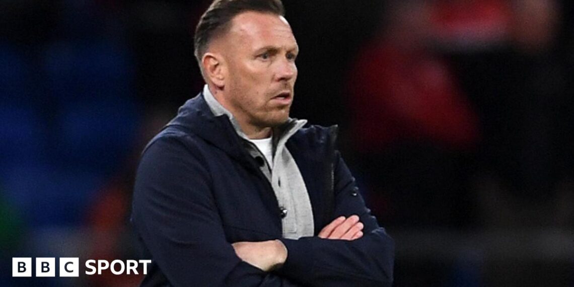 Craig Bellamy, in an open dark jacket with a grey top underneath, looks on with a focused stare during Wales' match against Northern Ireland
