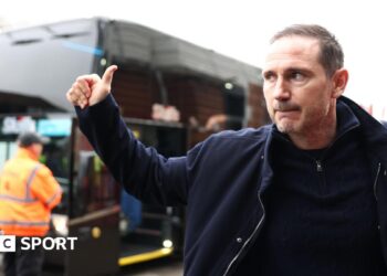 Frank Lampard gives a thumbs up in the direction of a bus outside a stadium.