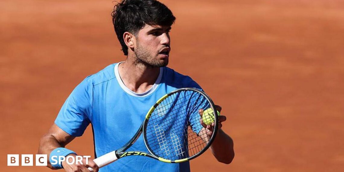 Carlos Alcaraz is wearing a blue t-shirt and holding a tennis ball to his racquet