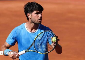 Carlos Alcaraz is wearing a blue t-shirt and holding a tennis ball to his racquet
