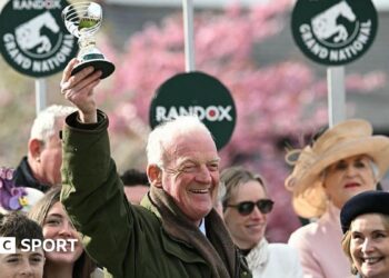 Willie Mullins smiling and holding a trophy in the air