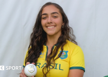 Laura Cardoso holding a white ball while wearing a Brazil cricket kit