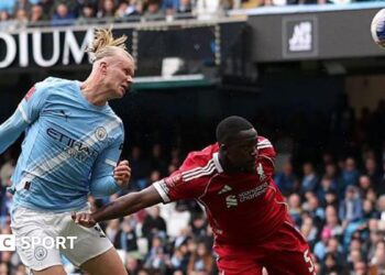 Erling Haaland, wearing Manchester City's light blue kit, celebrates his latest hat-trick by making a three-finger salute with his left hand
