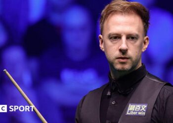 Judd Trump in black shirt and waistcoat carrying a snooker cue