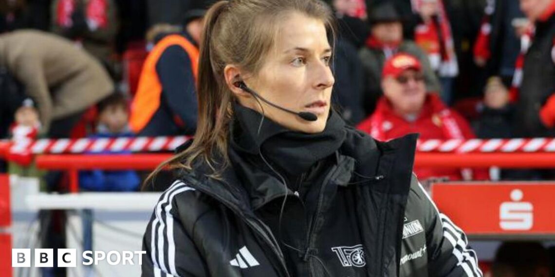 Marie-Louise Eta, wearing a black coat and top with a black microphone and headset in her right ear, on the touchline with fans in red and dark clothing in the background