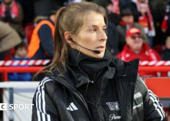 Marie-Louise Eta, wearing a black coat and top with a black microphone and headset in her right ear, on the touchline with fans in red and dark clothing in the background
