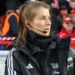 Marie-Louise Eta, wearing a black coat and top with a black microphone and headset in her right ear, on the touchline with fans in red and dark clothing in the background