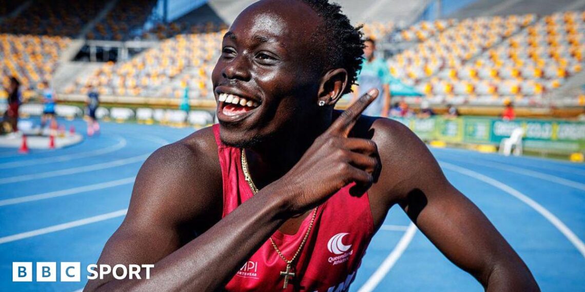 Gout Gout, wearing a red vest and a gold crucifix around his neck, points with his right hand across his body and smiles while crouching on a blue athletics track with white and yellow stadium seating in the background.