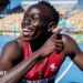 Gout Gout, wearing a red vest and a gold crucifix around his neck, points with his right hand across his body and smiles while crouching on a blue athletics track with white and yellow stadium seating in the background.