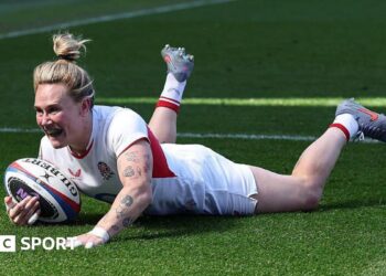 Meg Jones, wearing a white England shirt, shorts, and socks with her blonde hair tied up in a bin, grins as she dives over the tryline with the ball in her right hand