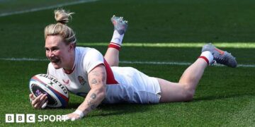Meg Jones, wearing a white England shirt, shorts, and socks with her blonde hair tied up in a bin, grins as she dives over the tryline with the ball in her right hand
