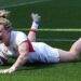 Meg Jones, wearing a white England shirt, shorts, and socks with her blonde hair tied up in a bin, grins as she dives over the tryline with the ball in her right hand