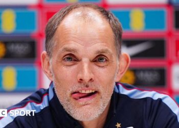 Head and shoulders view of England head coach Thomas Tuchel at a news conference, with his tongue poking out of his mouth