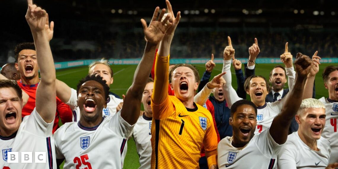 In a scene from Dear England, a team of footballers wearing the England men's team white and red kit celebrate on a football pitch, their goalkeeper, wearing yellow stands in the middle.