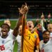 In a scene from Dear England, a team of footballers wearing the England men's team white and red kit celebrate on a football pitch, their goalkeeper, wearing yellow stands in the middle.