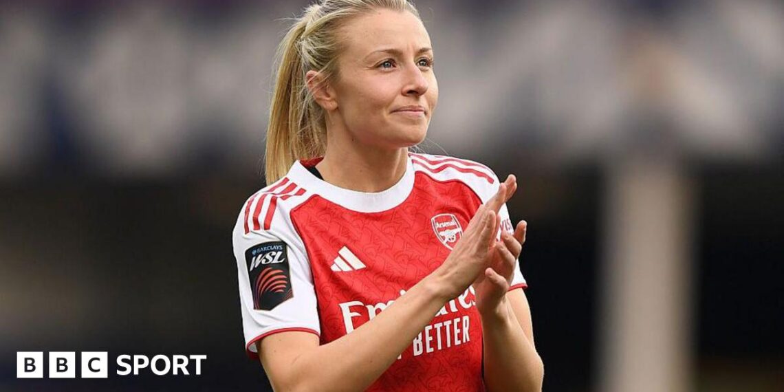 Leah Williamson, in a red Arsenal shirt with white sleeves and blonde hair tied back in a ponytail, looks in the direction of the crowd and applauds