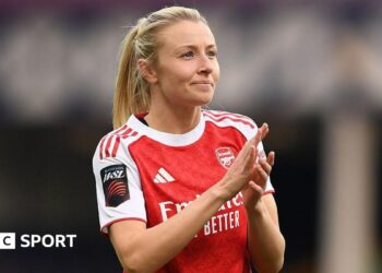 Leah Williamson, in a red Arsenal shirt with white sleeves and blonde hair tied back in a ponytail, looks in the direction of the crowd and applauds