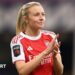 Leah Williamson, in a red Arsenal shirt with white sleeves and blonde hair tied back in a ponytail, looks in the direction of the crowd and applauds
