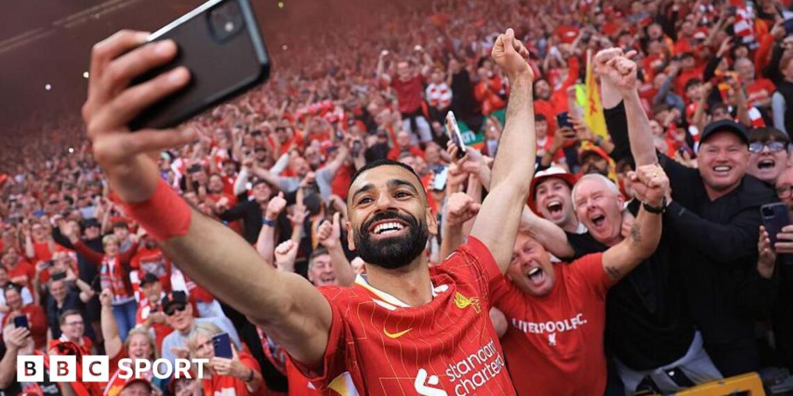 A grinning Mohamed Salah, in red club kit, takes a selfie with a mobile phone in front of excited Liverpool supporters