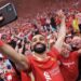 A grinning Mohamed Salah, in red club kit, takes a selfie with a mobile phone in front of excited Liverpool supporters
