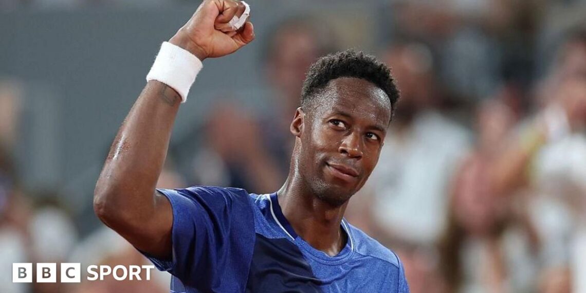 Novak Djokovic, in a pale yellow T-shirt, stands facing Gael Monfils, wearing a white T-shirt, rests his right arm on the Frenchman's shoulder