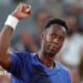 Novak Djokovic, in a pale yellow T-shirt, stands facing Gael Monfils, wearing a white T-shirt, rests his right arm on the Frenchman's shoulder