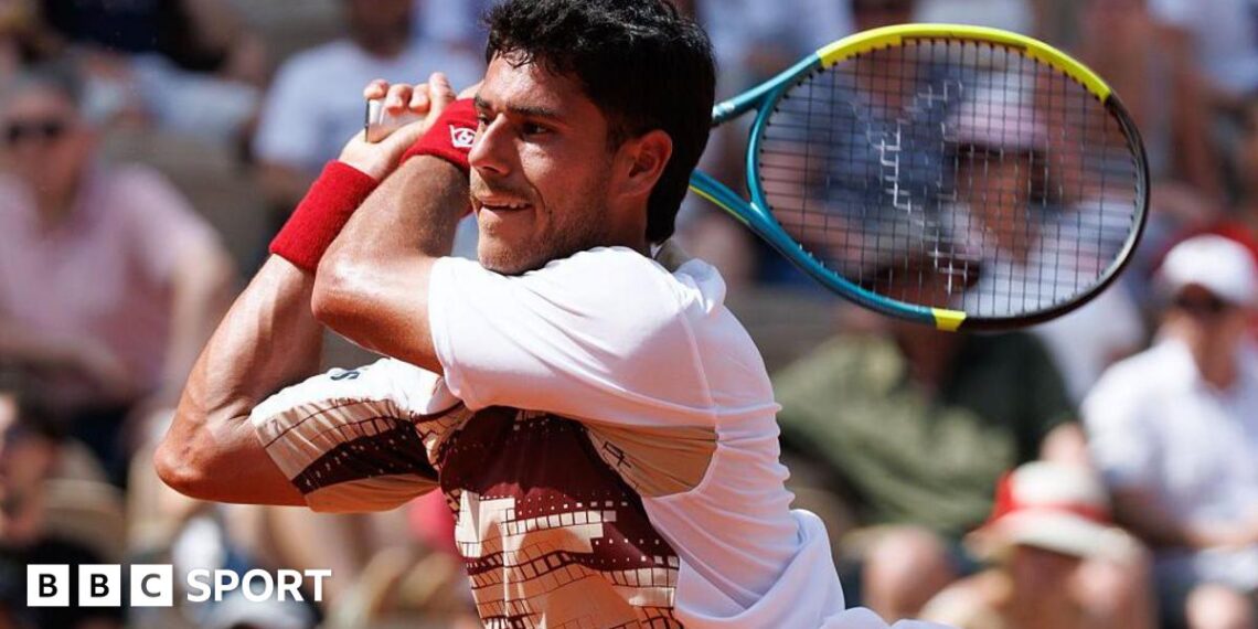 Adolfo Daniel Vallejo, wearing a white patterned t-shirt, plays a backhand with his yellow, blue and black racquet held in two hands over his right shoulder