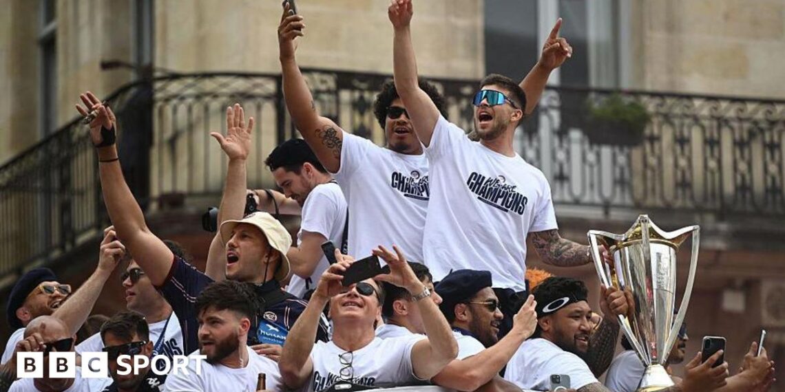 Bordeaux Begles players celebrate last year's Champions Cup victory at an event in the city, lifting the large trophy as a flag-waving crowd stand behind them. The players wear white T-shirts with the slogan WE ARE CHAMPIONS