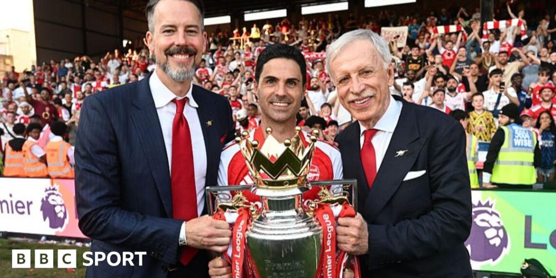 Josh Kroenke, Mikel Arteta and Stan Kroenke posed with the Premier League trophy, standing in front of a stand of Arsenal supporters who are largely dressed in red and white. Josh and Stan Kroenke wear dark suits, white shirts and a red tie each, while Arteta is in a red Arsenal shirt