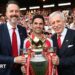 Josh Kroenke, Mikel Arteta and Stan Kroenke posed with the Premier League trophy, standing in front of a stand of Arsenal supporters who are largely dressed in red and white. Josh and Stan Kroenke wear dark suits, white shirts and a red tie each, while Arteta is in a red Arsenal shirt