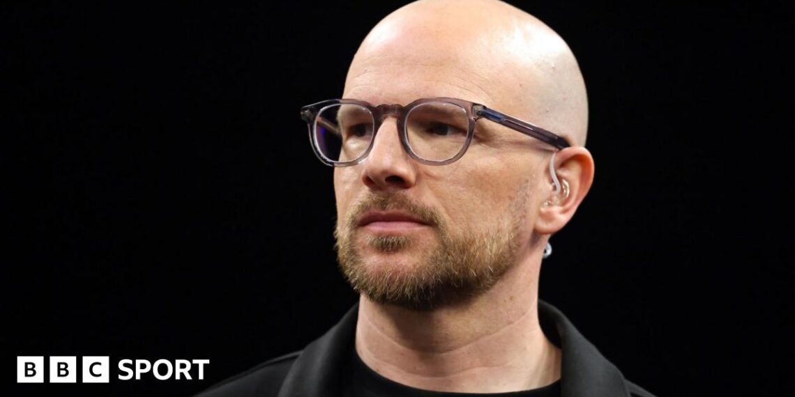 A close up of Mark Webster in dark shirt and jacket wearing spectacles and a TV earpiece
