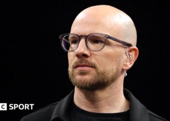 A close up of Mark Webster in dark shirt and jacket wearing spectacles and a TV earpiece