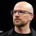 A close up of Mark Webster in dark shirt and jacket wearing spectacles and a TV earpiece