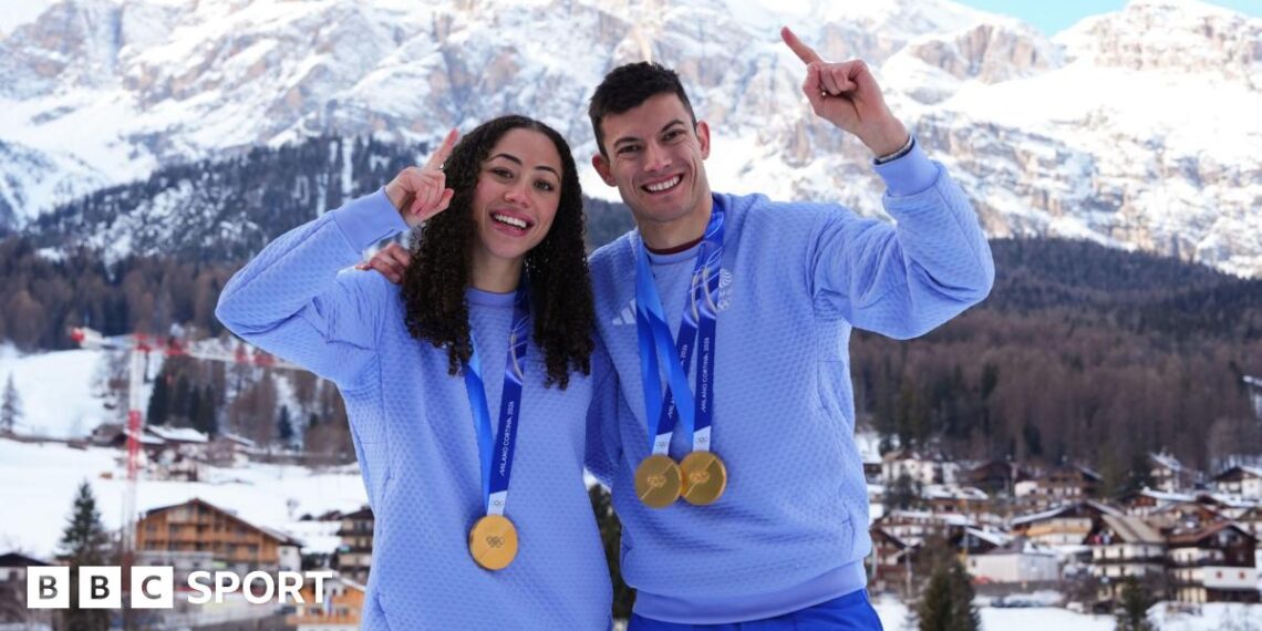 Tabby Stoecker (left) and Matt Weston stand with one arm around each other and the other in the air pointing their index fingers. They are both wearing Team GB kit and Winter Olympic gold medals around their necks. There are snow-capped moutains in the background