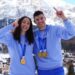 Tabby Stoecker (left) and Matt Weston stand with one arm around each other and the other in the air pointing their index fingers. They are both wearing Team GB kit and Winter Olympic gold medals around their necks. There are snow-capped moutains in the background