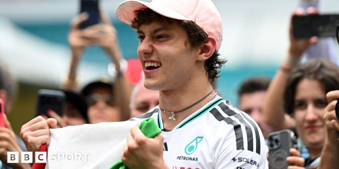 Kimi Antonelli celebrates his race win in Miami while holding the Italian flag. A crowd are gathered around him and taking photographs